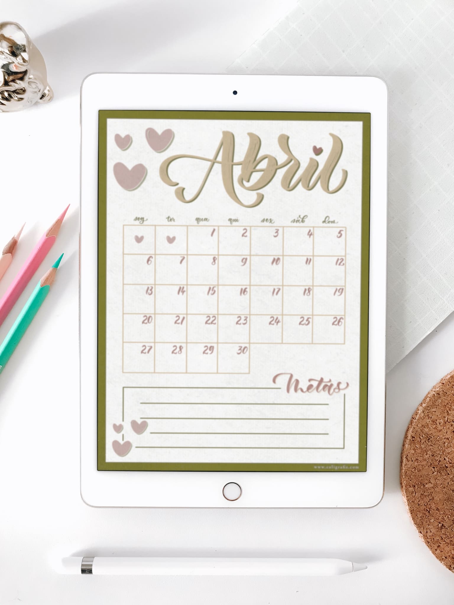 iPad com imagem de Planner digital do mês de abril sobre mesa com elementos decorativos ao redor